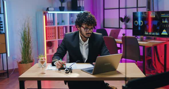 Indian Businessman Working on Laptop and Writing out Needed Information alt