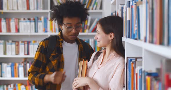 Young Multiethnic Couple Standing Against Bookshelf in College Library alt