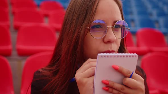 Young Woman in Glasses with Notepad Pen Sitting on Stadium Bleachers Alone alt