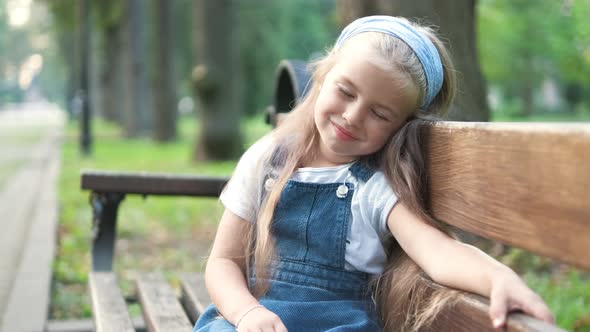 Small tired child girl sitting on a bench with closed eyes resting in summer park. alt
