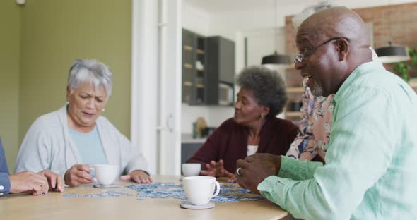 Group of happy diverse senior friends drinking coffee and doing puzzle at home alt