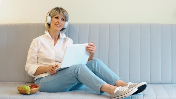 An Elderly Beautiful Slender Woman in Headphones and a Laptop in Her Hands Listens to Music with