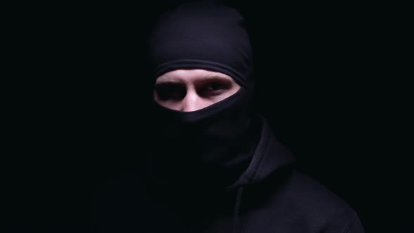 Criminal in Dark Balaclava Angrily Looking Into Camera Against Black Background alt