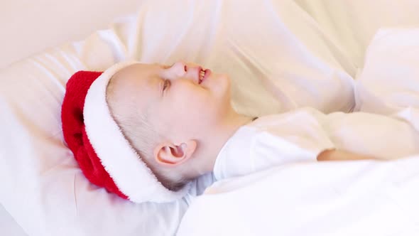 Child wakes up before Christmas. Little boy in Santa hat and pajamas lying in white bed.