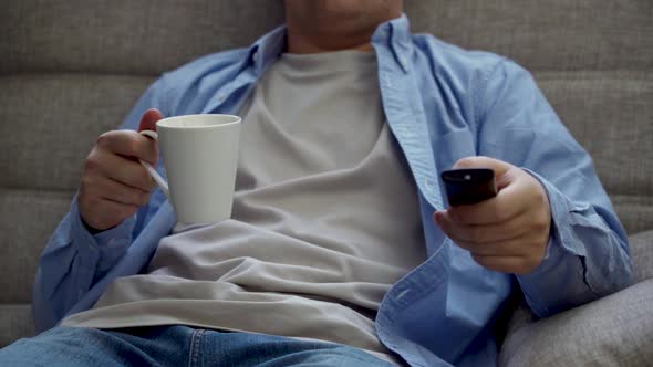 A Man 3040 Years Old Sits on the Sofa and Turns on the TV From the Remote Control alt