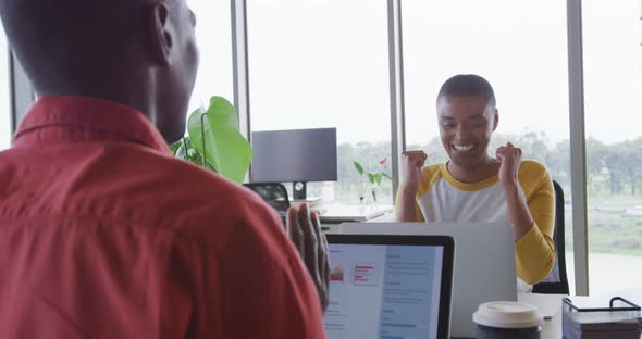 Happy african american creative businesswoman celebrating at desk in office with male colleague alt