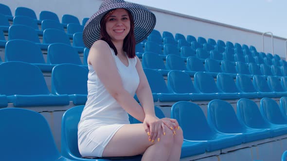 Young woman in white dress and elegant hat looks at camera, sitting on stadium bleachers alone. alt