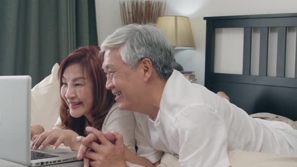 Asian senior couple using laptop after wake up, watching movie lying on bed in bedroom. alt