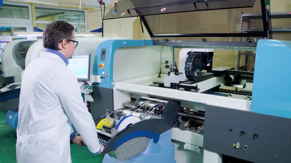 The Engineer in a White Robe and Glasses Works for the Surface Mount Technology Machine, PCB