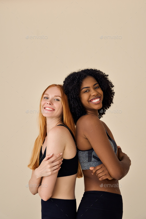 Two happy diverse fit women wearing sportswear standing on beige  background. Stock Photo by insta_photos