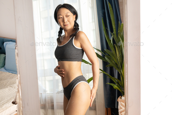 asian woman wearing underclothes has good body shape and looking her waist  in mirror. Stock Photo by nikolast1