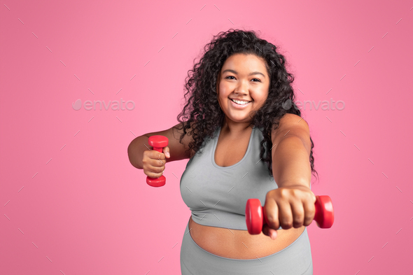 Cheerful excited black chubby lady in sportswear doing exercises with hand dumbbells, standing on