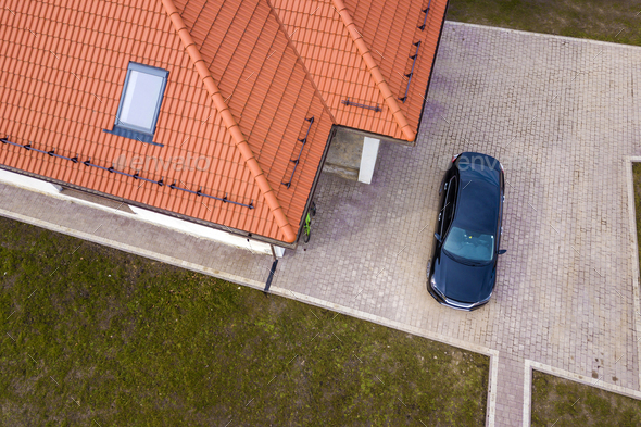 Aerial top view of house metal shingle roof with attic window and black car on paved yard.