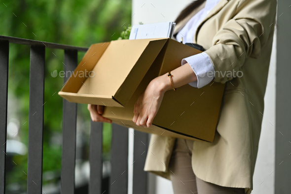Female employee holding cardboard box full of belongings leaving office after quitting job.