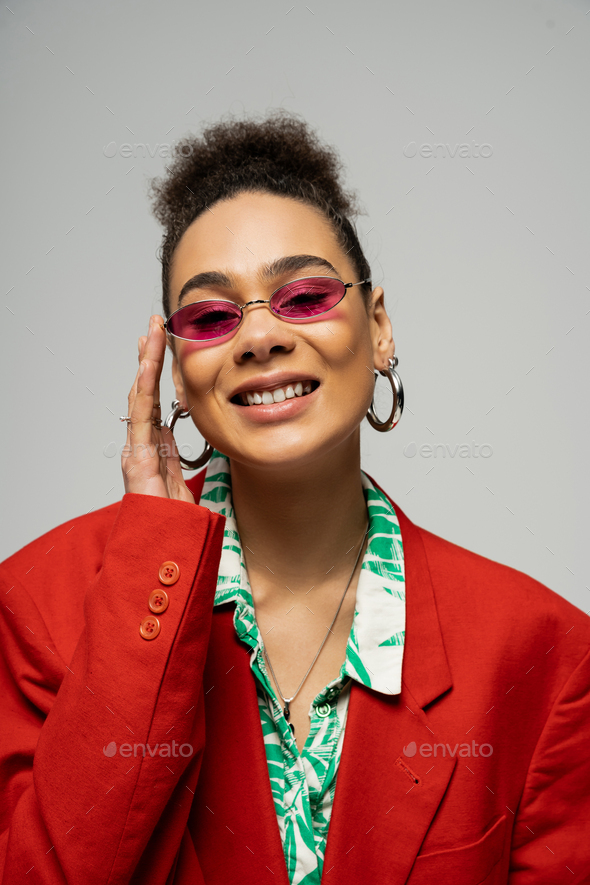 happy and stylish african american woman in trendy sunglasses and hoop earrings on grey backdrop