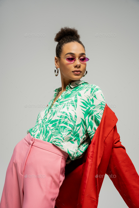 african american woman in sunglasses and trendy attire posing on grey backdrop, personal style