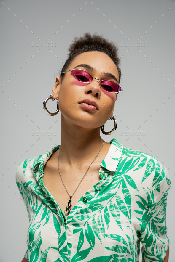 african american woman in pink sunglasses and hoop earrings looking at camera on grey backdrop
