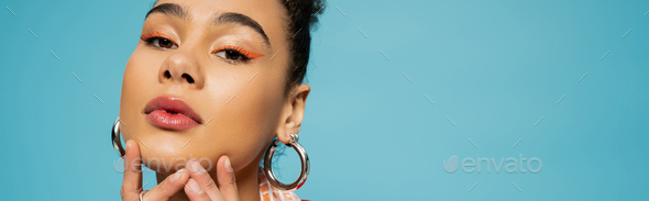 beautiful african american model in silver hoop earrings and rings looking at camera on blue, banner