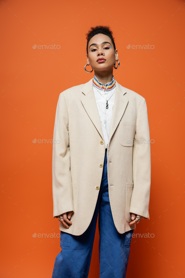 trendy young fashion model in beige blazer and jeans with hoop earrings posing on orange backdrop