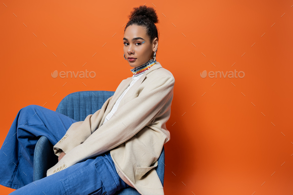 pretty fashion model in trendy attire with colourful makeup and hoop earrings posing on chair