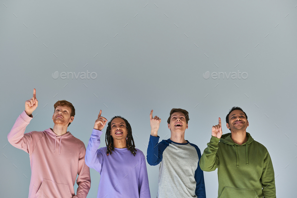 four joyful men in street wear looking up and pointing up on grey background, cultural diversity