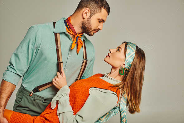 side view of retro style man in suspenders flirting with woman in orange dress on grey, hip couple