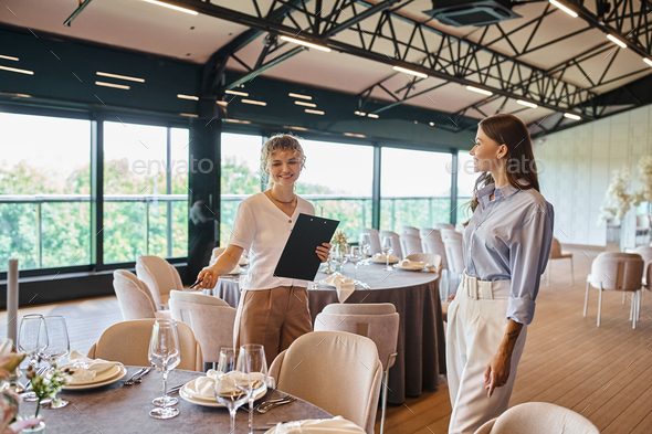 event organizer with clipboard pointing at table with festive setting near woman in banquet hall