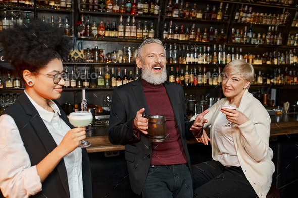 cheerful bearded man laughing near multiethnic women with cocktail glasses in bar, after work party