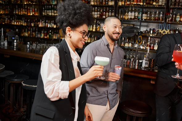 asian man and african american woman holding cocktails and smiling during after work party in bar