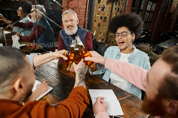 overjoyed multiethnic workmates clinking beer bottles while resting in pub after work, party time