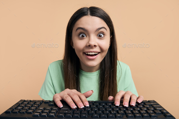 Surprised beautiful teen girl with WOW emotion looking at camera, typing text on laptop keyboard