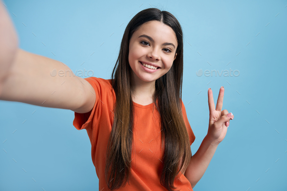 Self-portrait of pretty teen girl showing peace sign, smiling looking at camera on her mobile phone