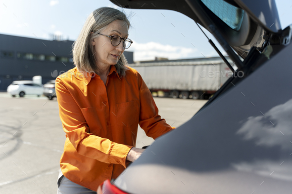 Close-up elegant senior woman driver opening the car trunk, standing in a parking lot outdoors