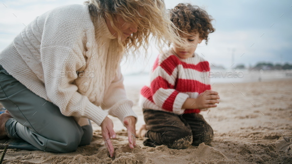 Family building sand castle on cool beach. Single mom playing little curly son