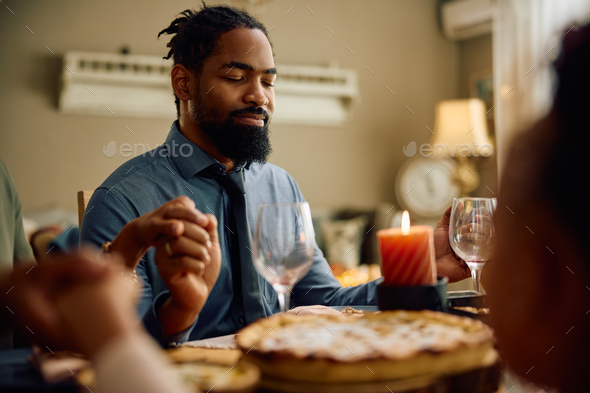 Black man praying with his family during Thanksgiving meal at dining table.