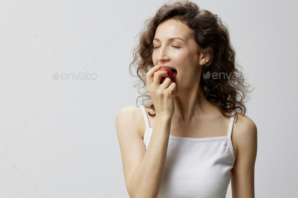Enjoyed cheerful lovely curly beautiful woman in basic white t-shirt enjoy fresh apple fruit posing