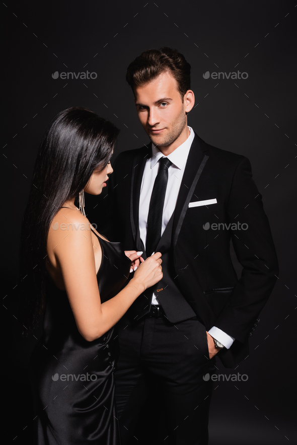 sensual brunette woman unbuttoning vest of elegant man standing with hand in pocket on black