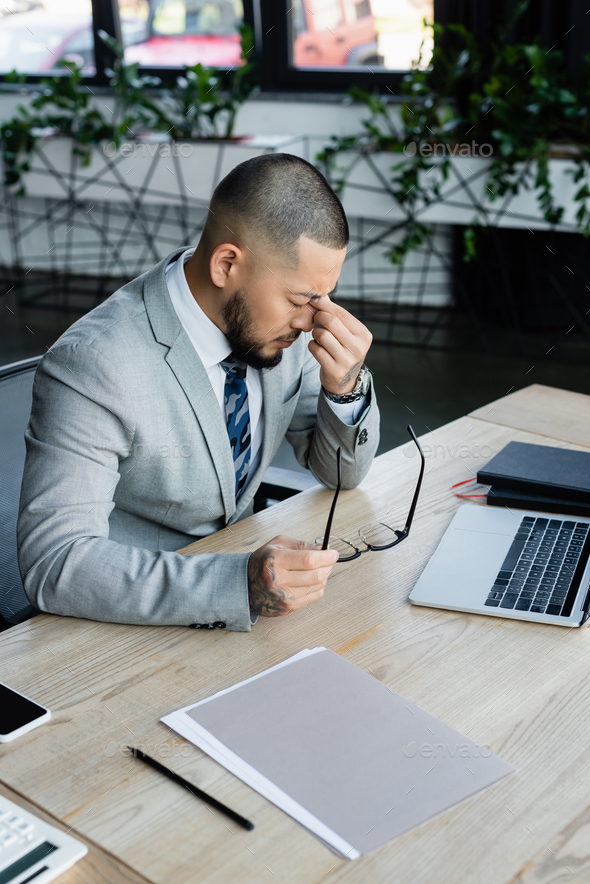 tired asian businessman holding eyeglasses and rubbing eyes near laptop