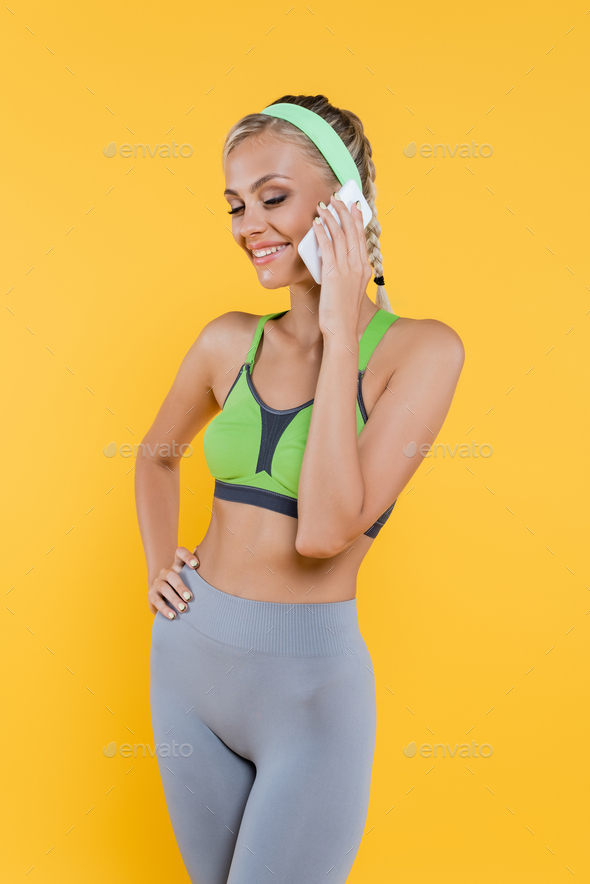 sportive woman in green sports bra and grey leggings standing with hand on hip and talking on