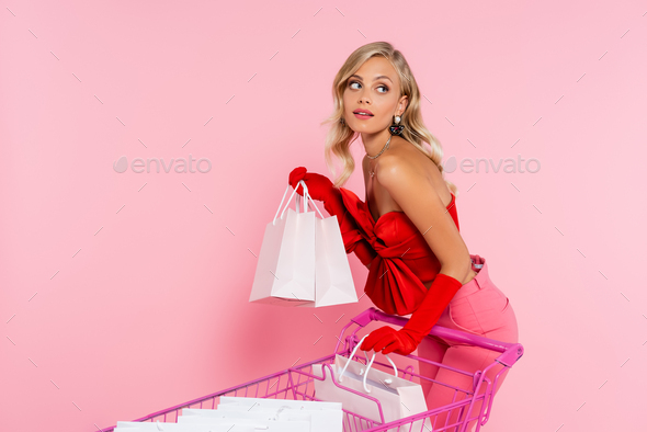 fashionable blonde woman in red gloves posing near shopping cart with purchases on pink