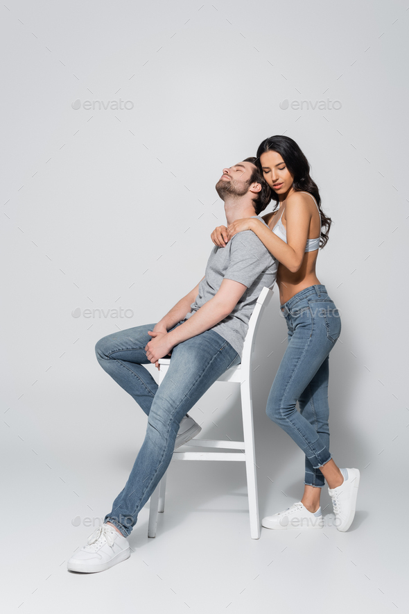 young and brunette woman in bra and jeans looking away while sitting on  grey Stock Photo by LightFieldStudios