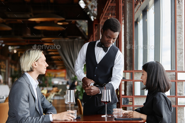 Server showing label on red wine bottle to couple in restaurant