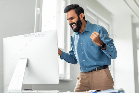 Business success. Overjoyed indian businessman shaking fists while looking at computer monitor in