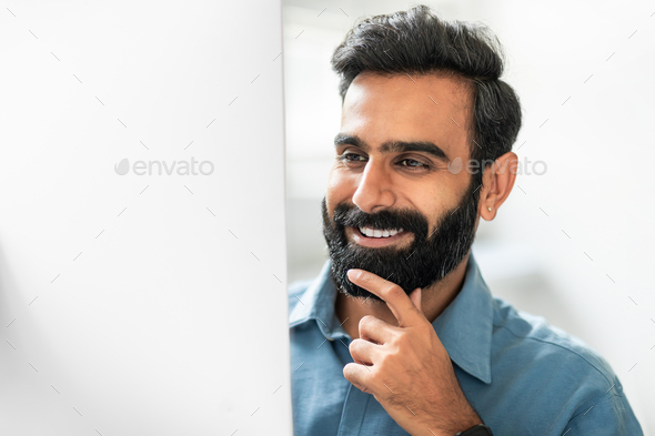 Happy indian businessman working on computer in office, looking at monitor and smiling, touching