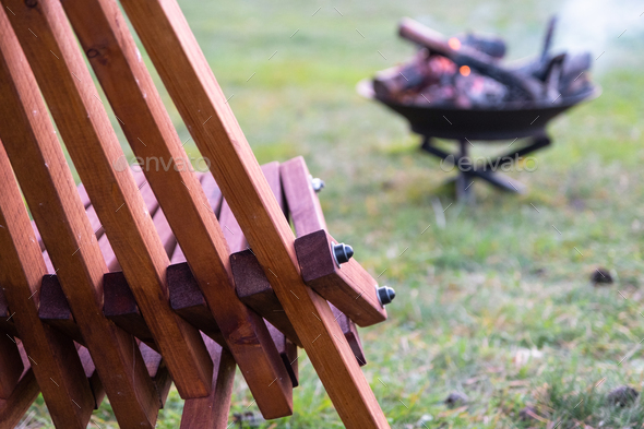 Metal fire bowl with burning wood in the yard and garden rack deck chairs. Safe decorative fire pit,