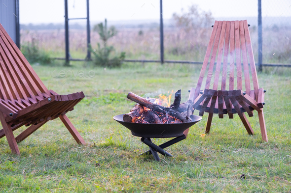 Metal fire bowl with burning wood in the yard and garden rack deck chairs. Safe decorative fire pit,