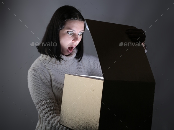Young Hungarian woman in a sweater opening a mystery box with a shocked expression
