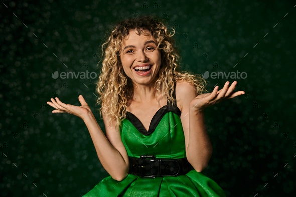 joyous curly woman in new year elf costume standing under falling snow on dark green background