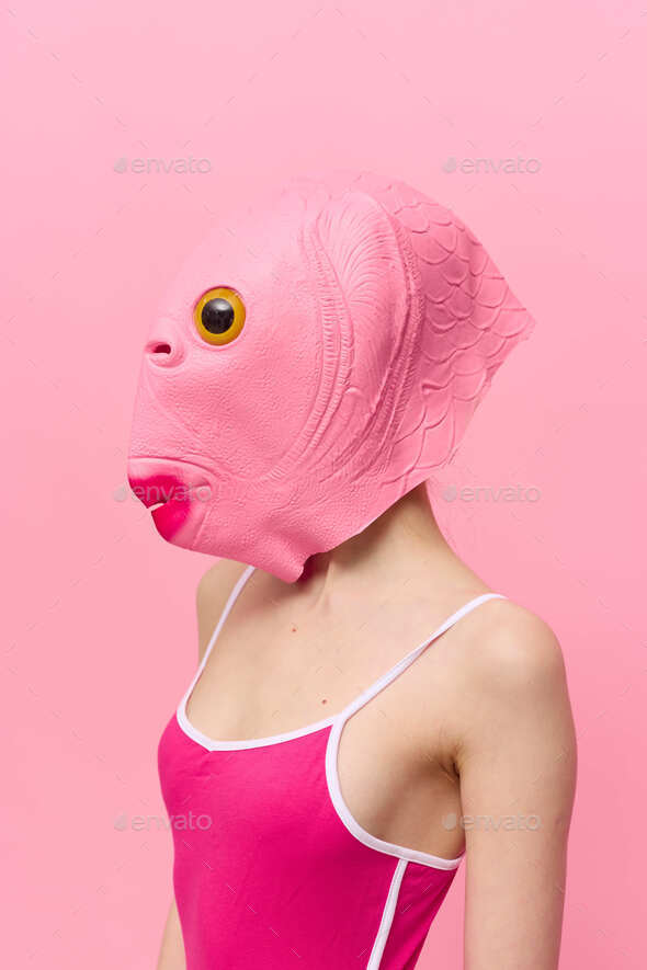 A woman in a pink fish head mask stands in profile against a pink