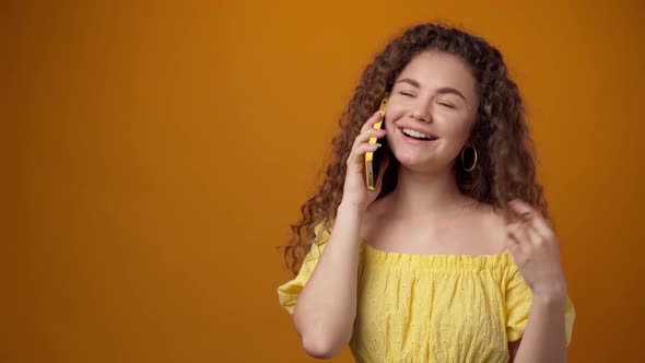 Young Curly Woman Talking on the Phone and Smiling Against Yellow Backrgound